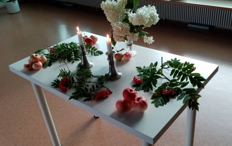 Valkoisella pöydällä valkoisia syyshortensian kukkia lasimaljakossa, kaksi palavaa kynttilää tinaisissa kynttilänjaloissa, pihlajan oksia, joissa punaisia pihlajanmarjoja ja vielä vihreitä lehtiä; ja kolme pientä kekoa, joissa punaisia kaneliomenoita; pöytä on sisällä Hauhon seurakuntatalon juhlasalissa